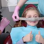 Girl in dental chair with nitrous oxide mask on nose giving 2 thumbs up